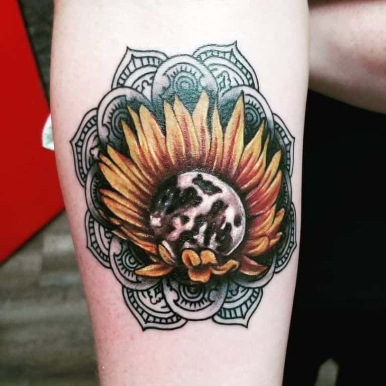 Bold sunflower tattoo on forearm with vibrant yellow petals, a moon-like center, and intricate mandala patterns in the background.