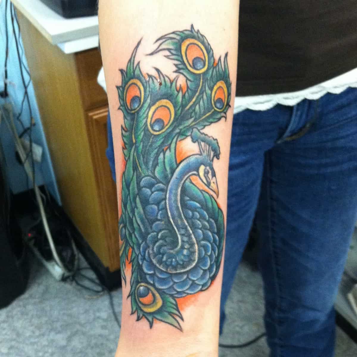 Forearm tattoo of a blue peacock with vibrant tail feathers in green and orange highlights