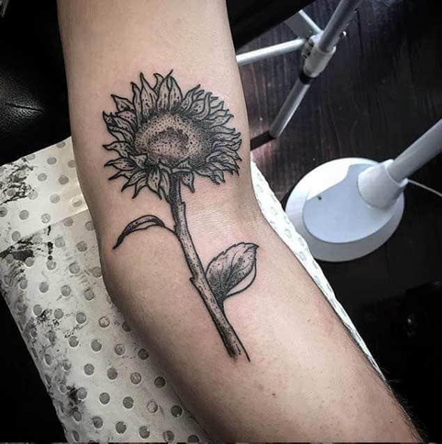 Black and grey sunflower tattoo on forearm with bold shading, fine line details, and a slightly wilted stem for a unique, artistic look.