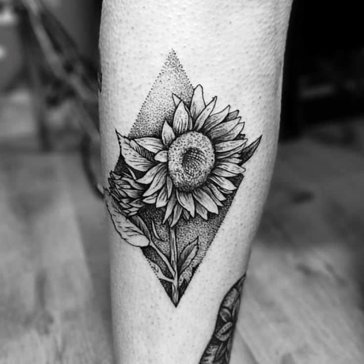 Black and grey sunflower tattoo on leg with fine line details, dotwork shading, and a geometric diamond-shaped background for depth.