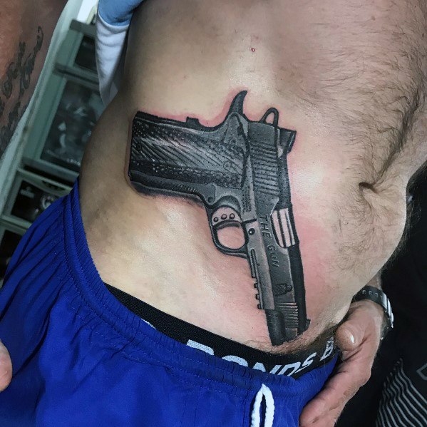Realistic black and gray tattoo of a 1911 pistol on the side of the torso, designed to appear as if the gun is tucked into the waistband.
