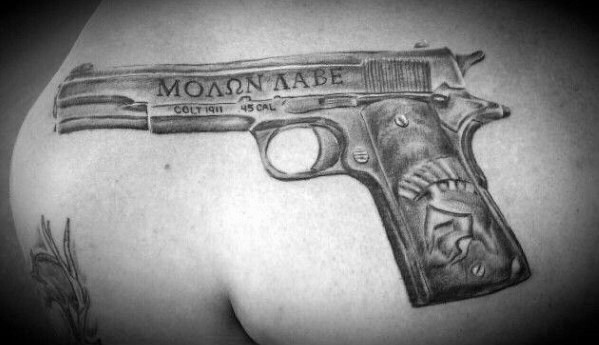 Black and gray tattoo of a Colt 1911 pistol on the upper back, featuring "ΜΟΛΩΝ ΛΑΒΕ" engraved on the slide and a Spartan helmet on the grip.