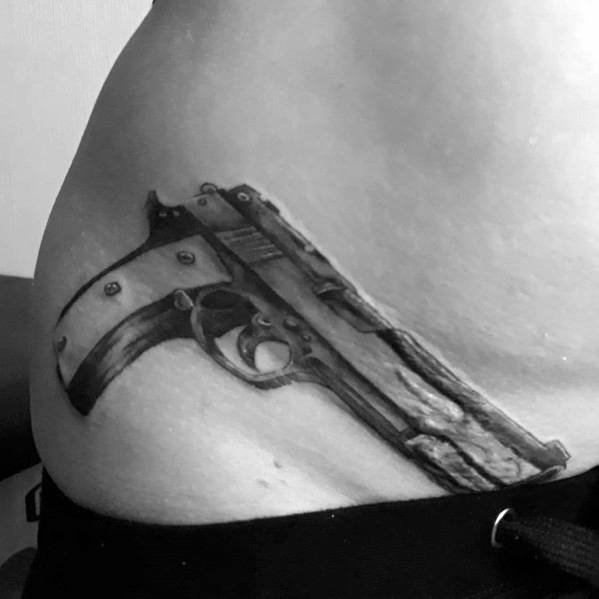 Realistic black and gray tattoo of a pistol on the waist, designed to appear as if it is tucked into the skin for a three-dimensional effect.