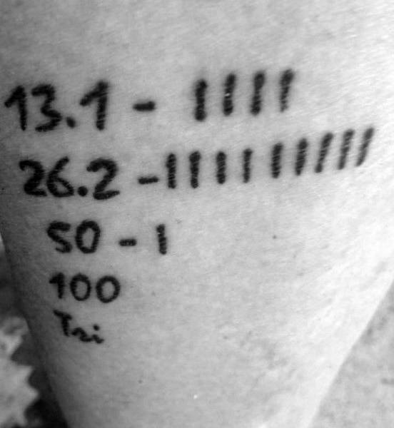 Minimalist tally-style tattoo listing race distances—13.1, 26.2, 50, 100—each marked with completed runs as tally marks.