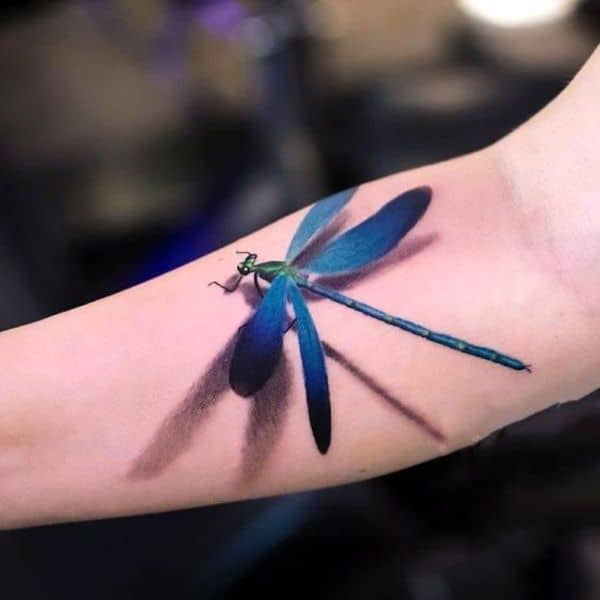 Realistic blue dragonfly tattoo on the inner arm, featuring a 3D effect with detailed shading and a lifelike shadow.