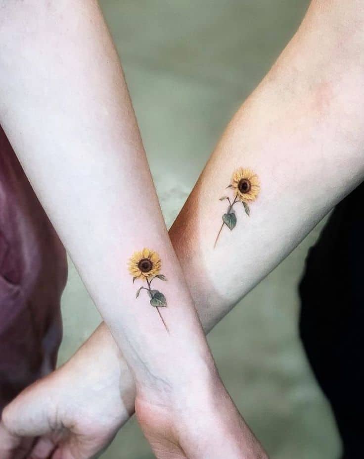 Matching minimalist sunflower tattoos on wrists, featuring fine details, soft yellow petals, and delicate green leaves for a subtle floral design.