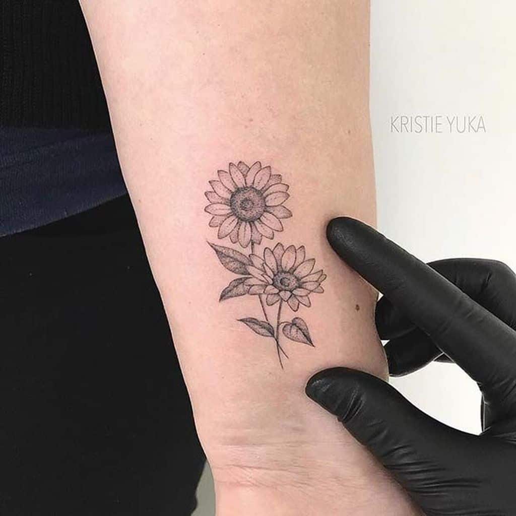 Small black ink tattoo of two sunflowers on the forearm, featuring fine line details and soft shading for a delicate floral design.