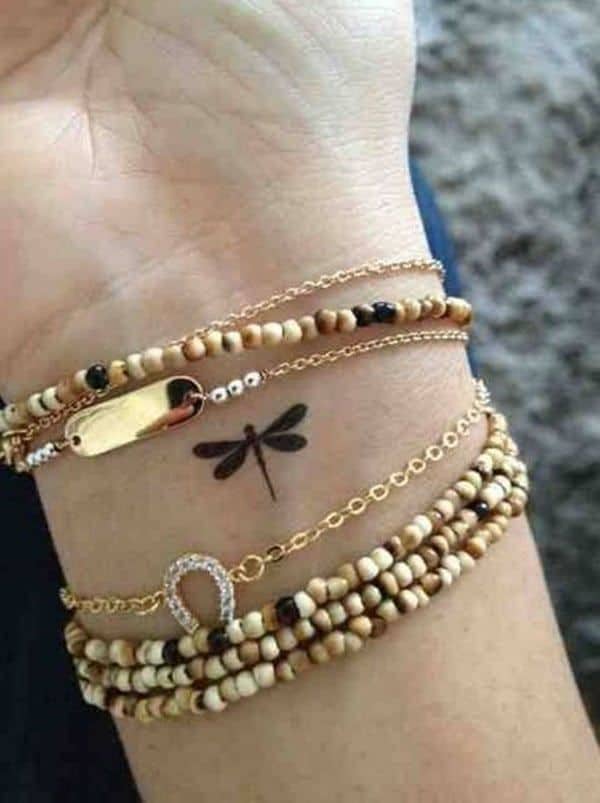Small black dragonfly tattoo on the wrist, surrounded by gold and beaded bracelets. The minimalist design adds a stylish touch.
