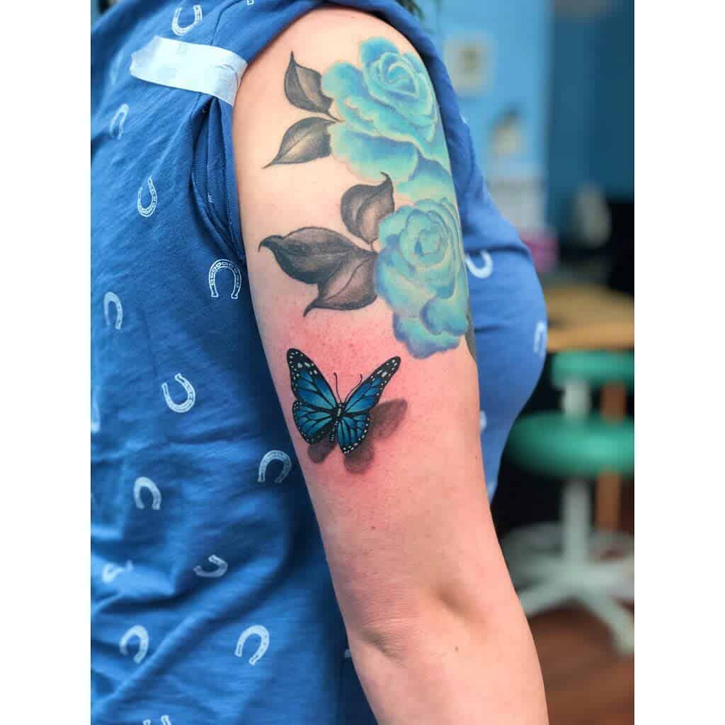 Person's arm adorned with tattoos of blue roses intertwined with a lifelike blue butterfly