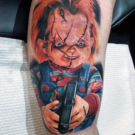 Chucky horror tattoo with vivid colors, showcasing his sinister grin, stitched face, and a gun in his hands for a menacing look.