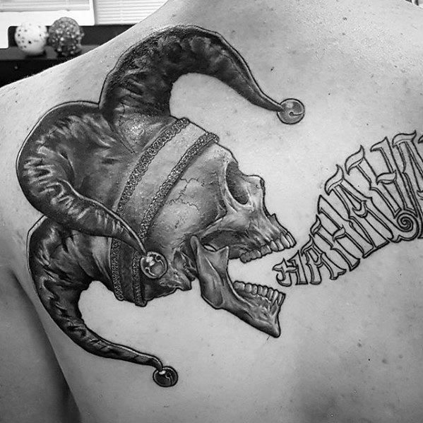 A detailed black and gray tattoo of a jester skull with a jester hat and stylized lettering, placed on the upper back.
