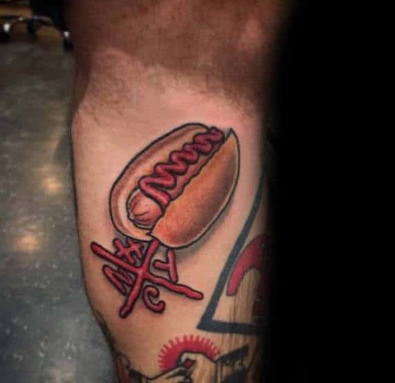 Hot dog tattoo featuring a stylized hot dog with red mustard on the upper arm, paired with playful cross details.