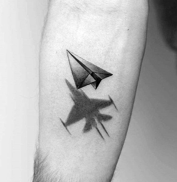 Paper airplane tattoo casting a jet fighter shadow, symbolizing dreams, ambition, and transformation in a bold black ink design.