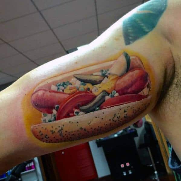 Realistic hot dog tattoo featuring a detailed sausage with toppings like tomatoes, onions, and mustard on a poppy seed bun.