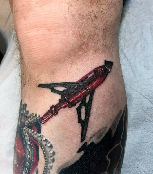 Realistic broadhead arrow tattoo with 3D effect, placed near the knee, appearing as if embedded in the skin.