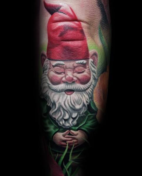 Gnome tattoo with a red hat, white beard, and green outfit, sitting peacefully with hands clasped, surrounded by greenery.