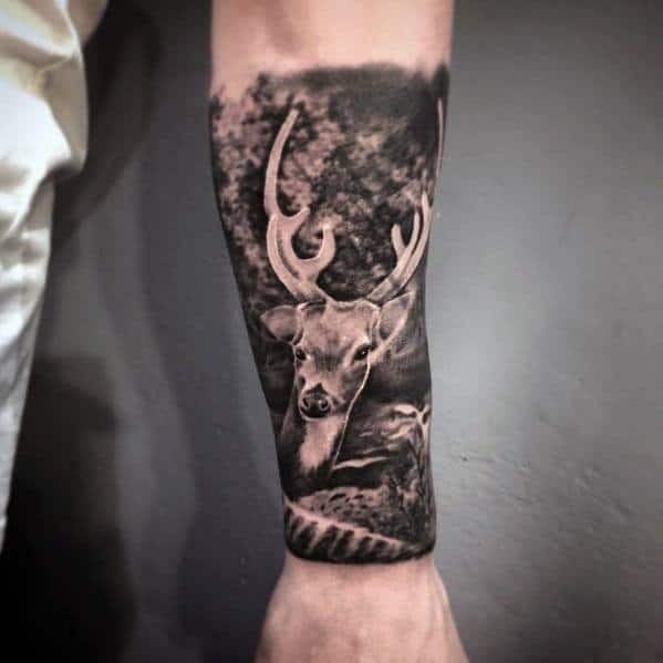 Realistic black and grey elk tattoo on a man's forearm, featuring intricate shading and a detailed forest background.