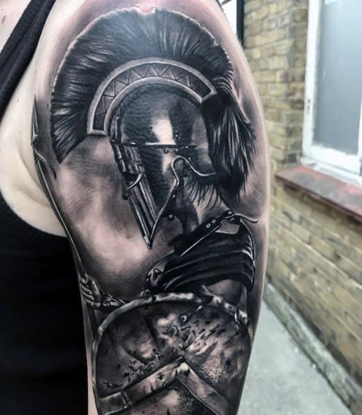 Realistic black and grey upper arm tattoo of a Spartan warrior in profile, featuring a plumed helmet, detailed armor, and weathered shield in battle.