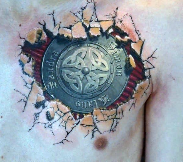Realistic chest tattoo of a Celtic shield beneath torn skin, revealing cracked flesh and muscle with engraved text encircling the emblem.