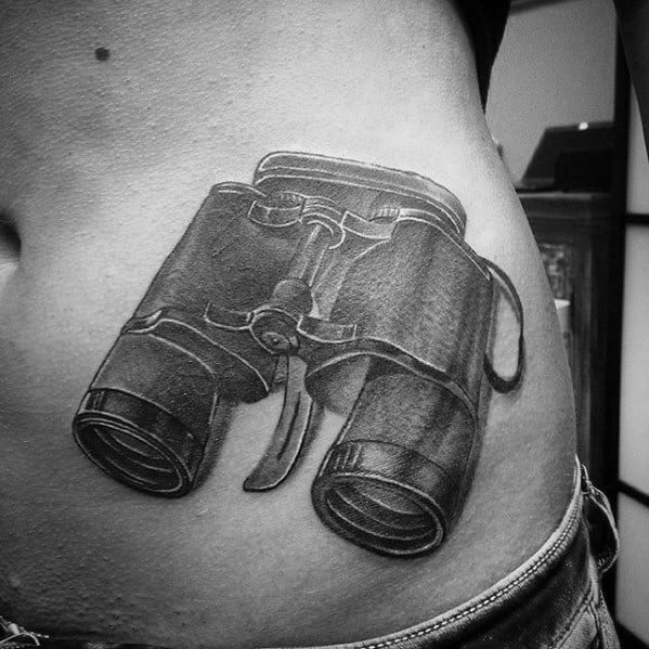 Realistic black and grey binoculars tattoo on the side of the torso, featuring fine shading and intricate details for a 3D effect.