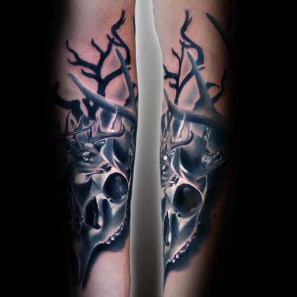 Realistic deer skull tattoo with large antlers and dark tree branches, covering both forearms in a dramatic black and gray style.