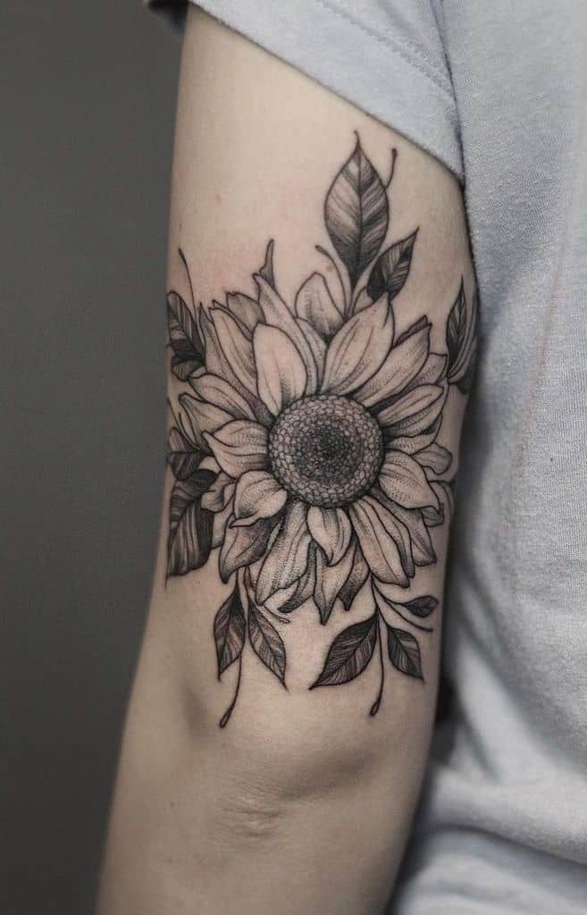 Black and grey sunflower tattoo on upper arm with intricate shading, fine line details, and surrounding leaves for a bold floral design.
