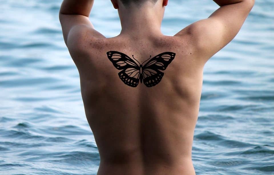 Bold black butterfly tattoo centered on the upper back, featuring symmetrical wings with intricate cut-out details against a waterfront backdrop.