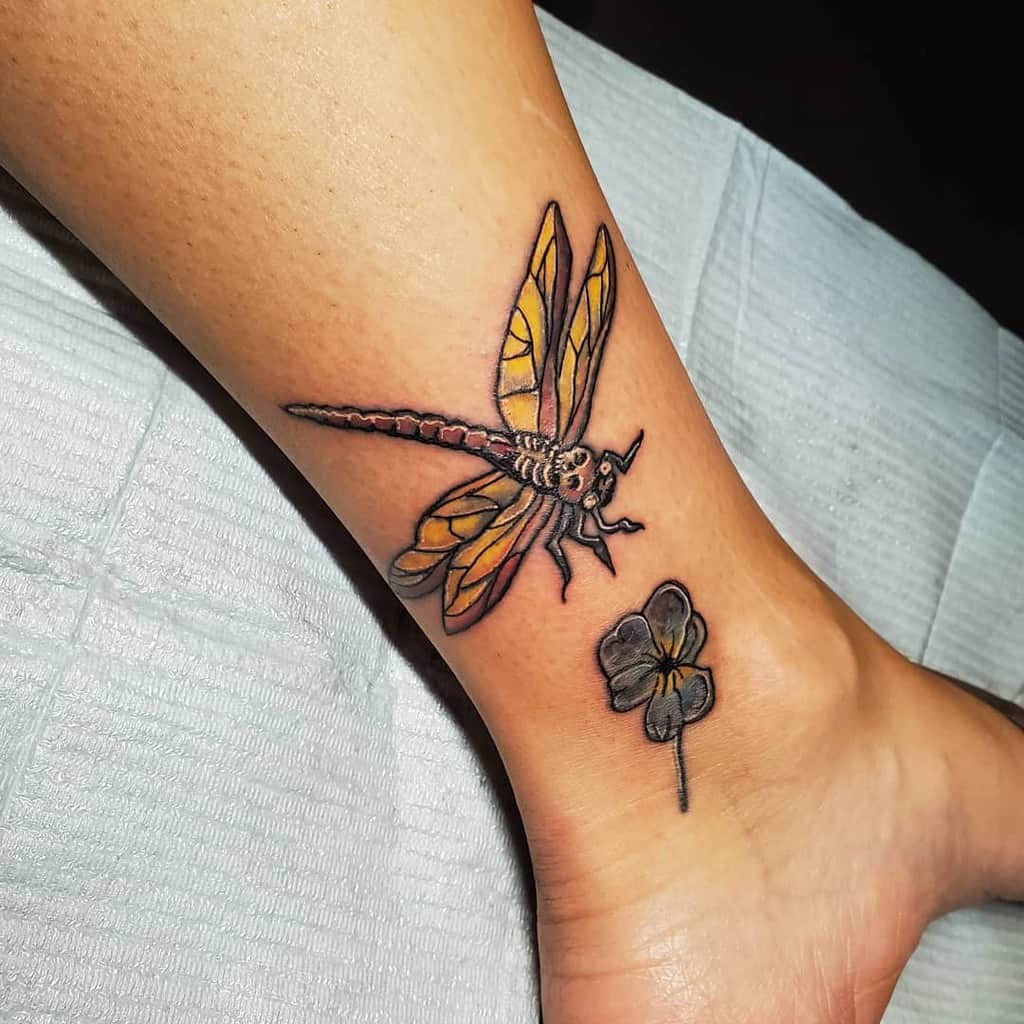 Realistic dragonfly tattoo with yellow-orange wings on the ankle, accompanied by a shaded four-leaf clover for a symbolic touch.