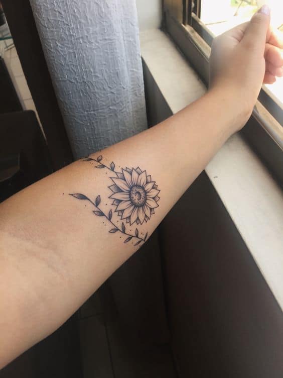 Delicate black ink sunflower tattoo on forearm with fine line details, leafy accents, and subtle shading for an elegant look.