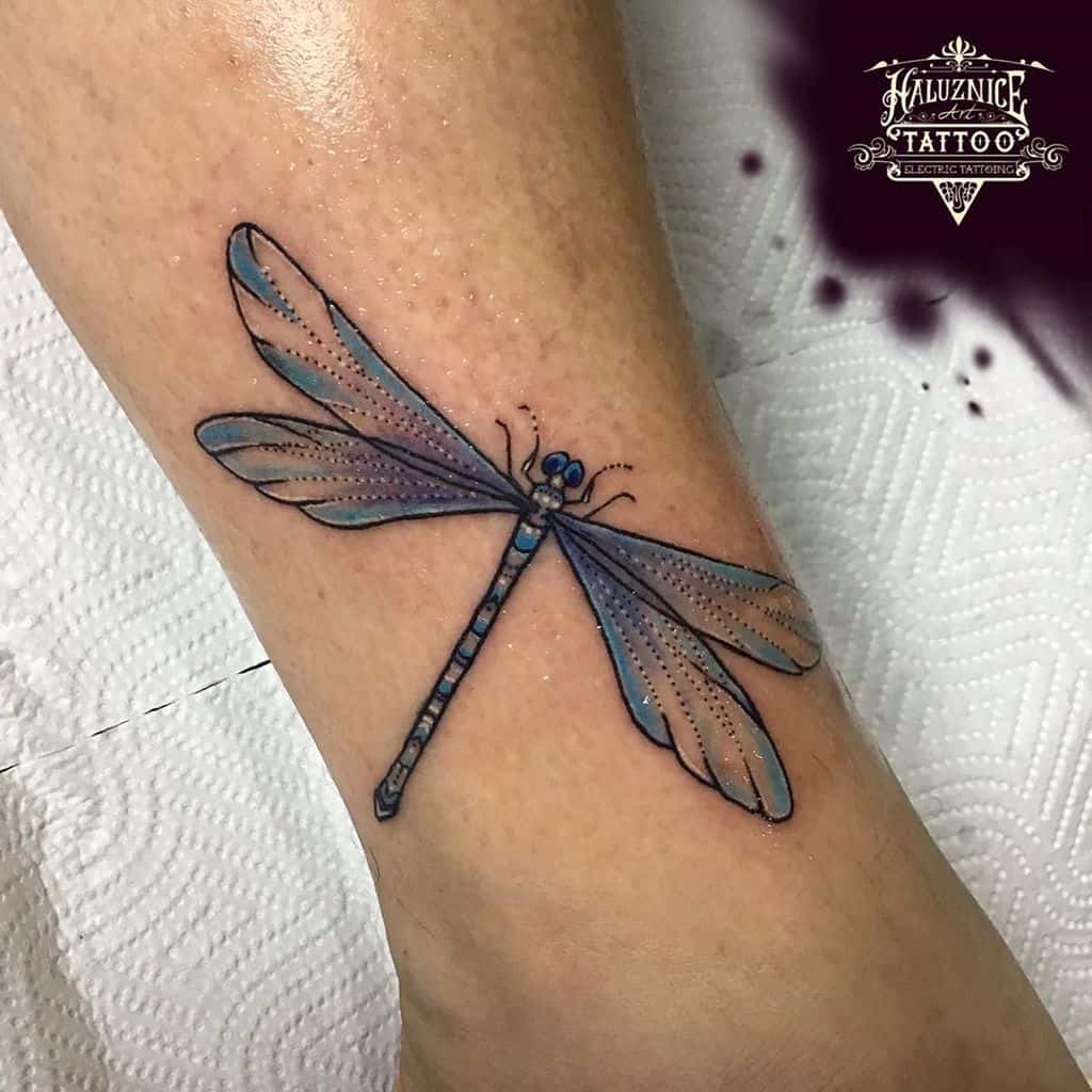 Colorful dragonfly tattoo on the ankle with blue and purple shading. The delicate wings and detailed body add a lifelike effect.