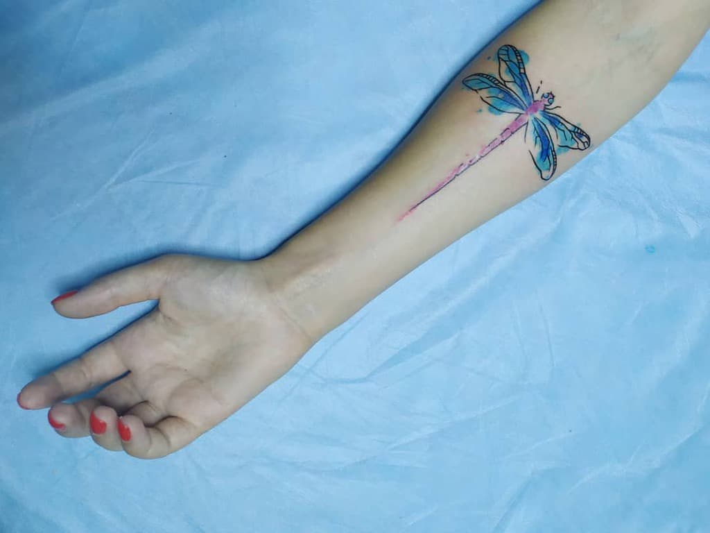 Elegant watercolor dragonfly tattoo on the forearm, featuring delicate blue wings and a flowing pink body for a soft, artistic look.
