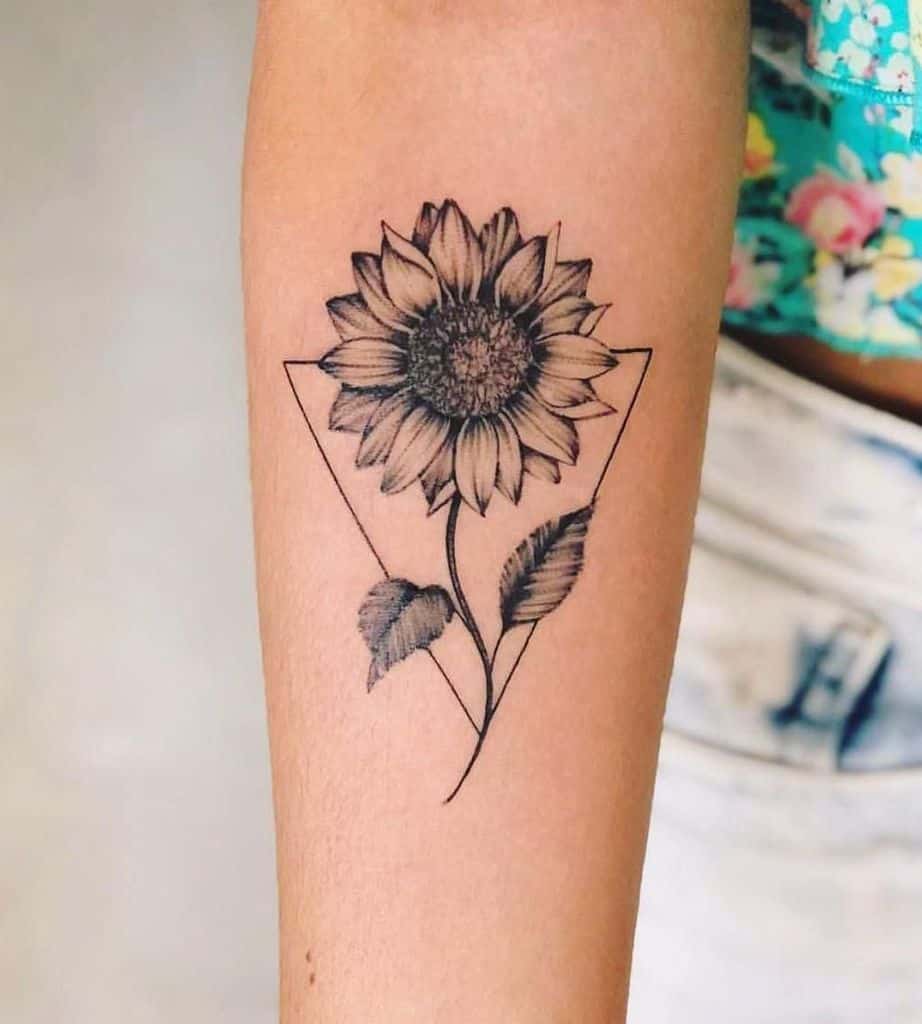 Minimalist black and grey sunflower tattoo on forearm, featuring fine line details, soft shading, and a geometric triangle frame.