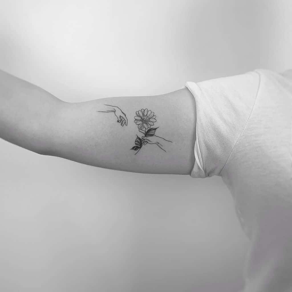 Minimalist sunflower tattoo on inner arm, featuring two hands reaching for the flower in delicate line art. Black ink design.