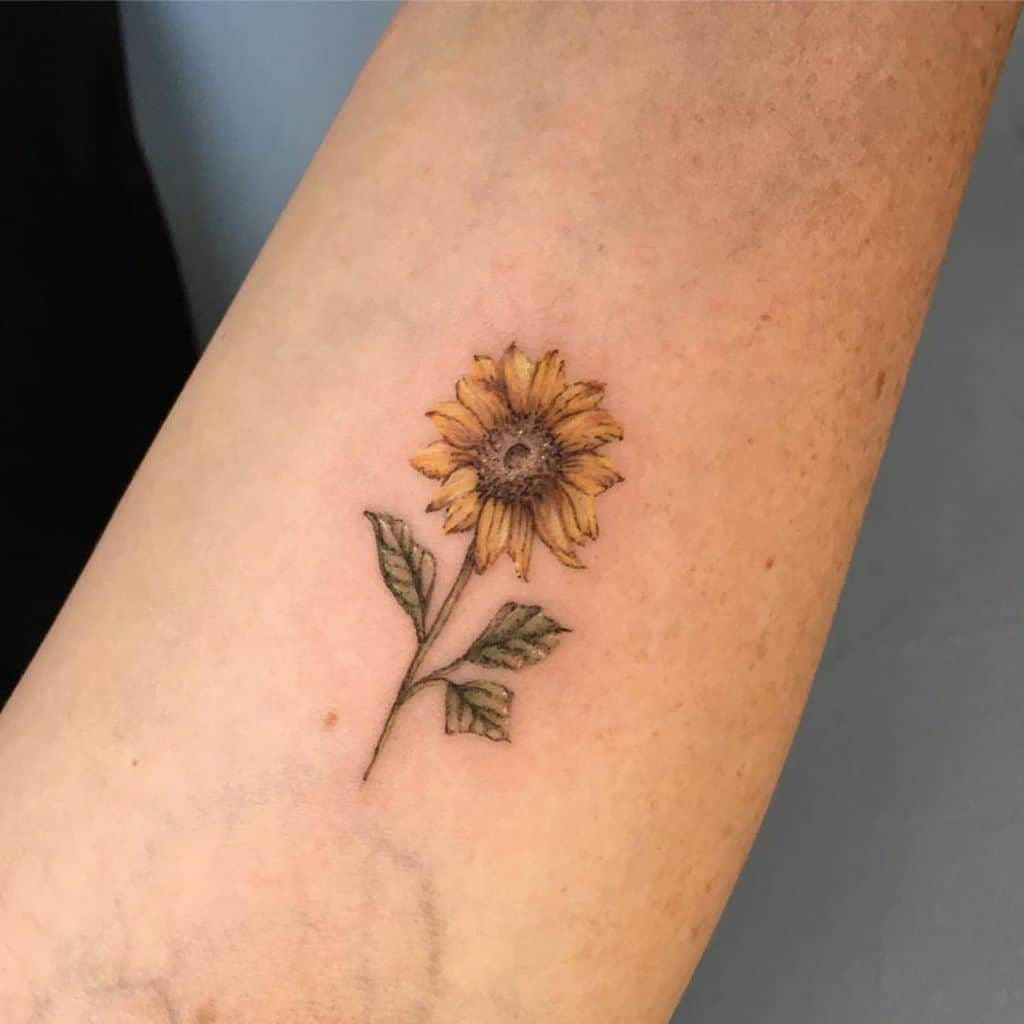 Minimalist sunflower tattoo on forearm with fine details, soft yellow petals, green leaves, and a delicate stem for a subtle floral design.