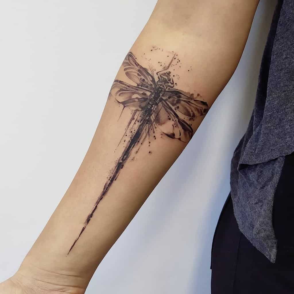 Bold black ink dragonfly tattoo on the forearm, featuring an abstract watercolor design with splashes and a long, flowing tail effect.