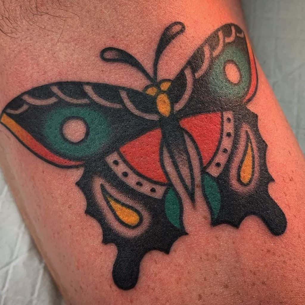 Bold neo-traditional butterfly tattoo featuring vibrant red, green, yellow, and black hues with thick outlines and intricate wing details.