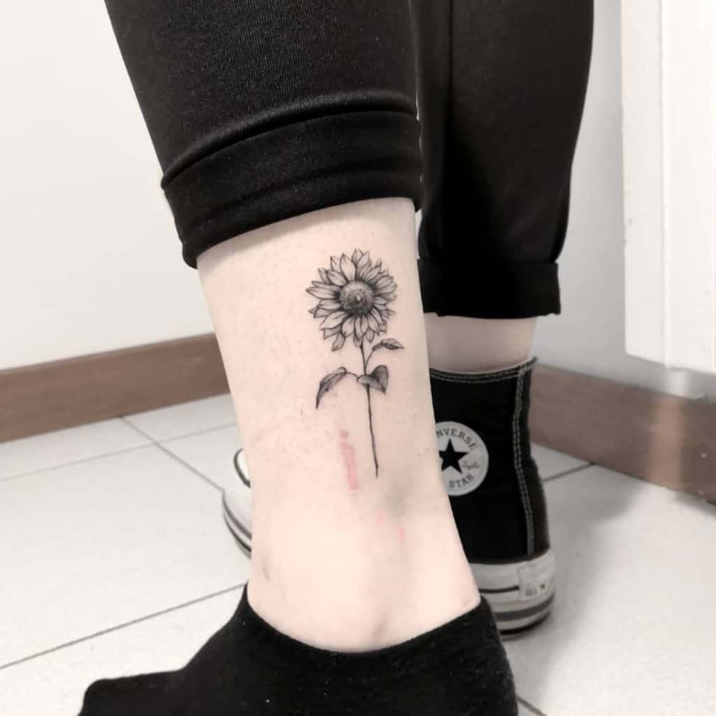 Small black ink sunflower tattoo on ankle with fine line detailing and a delicate stem, creating a minimalist yet elegant design.