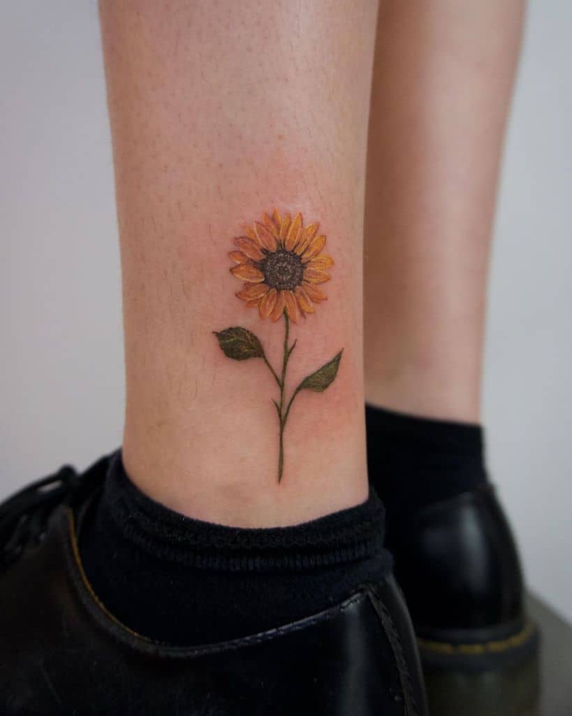 Small, delicate sunflower tattoo on the ankle with fine details, featuring yellow petals, a brown center, and green leaves for a realistic look.