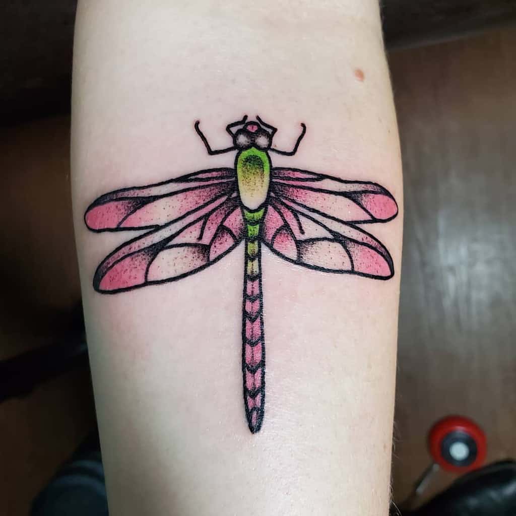 Colorful dragonfly tattoo on the forearm with pink wings and green accents, outlined in bold black for a vibrant look.