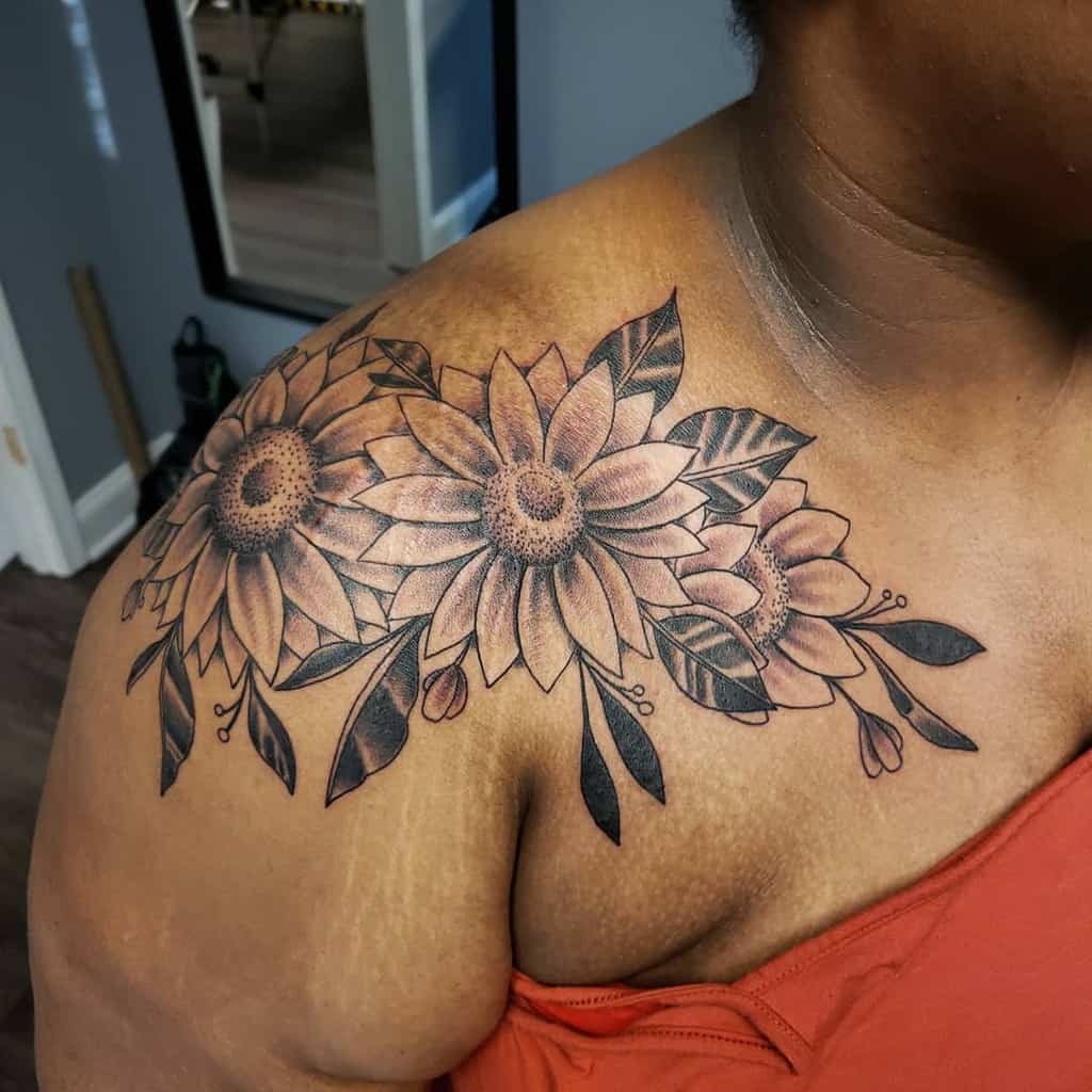 Black and grey sunflower tattoo on shoulder featuring three large sunflowers with fine shading, leaves, and floral accents for a bold design.