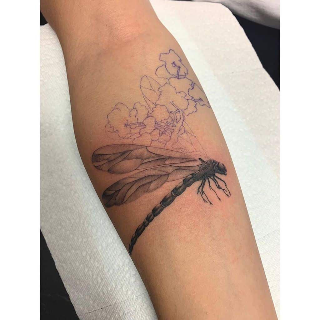 Black and grey dragonfly tattoo on the forearm with fine shading. Partially outlined flowers in the background add an artistic touch.