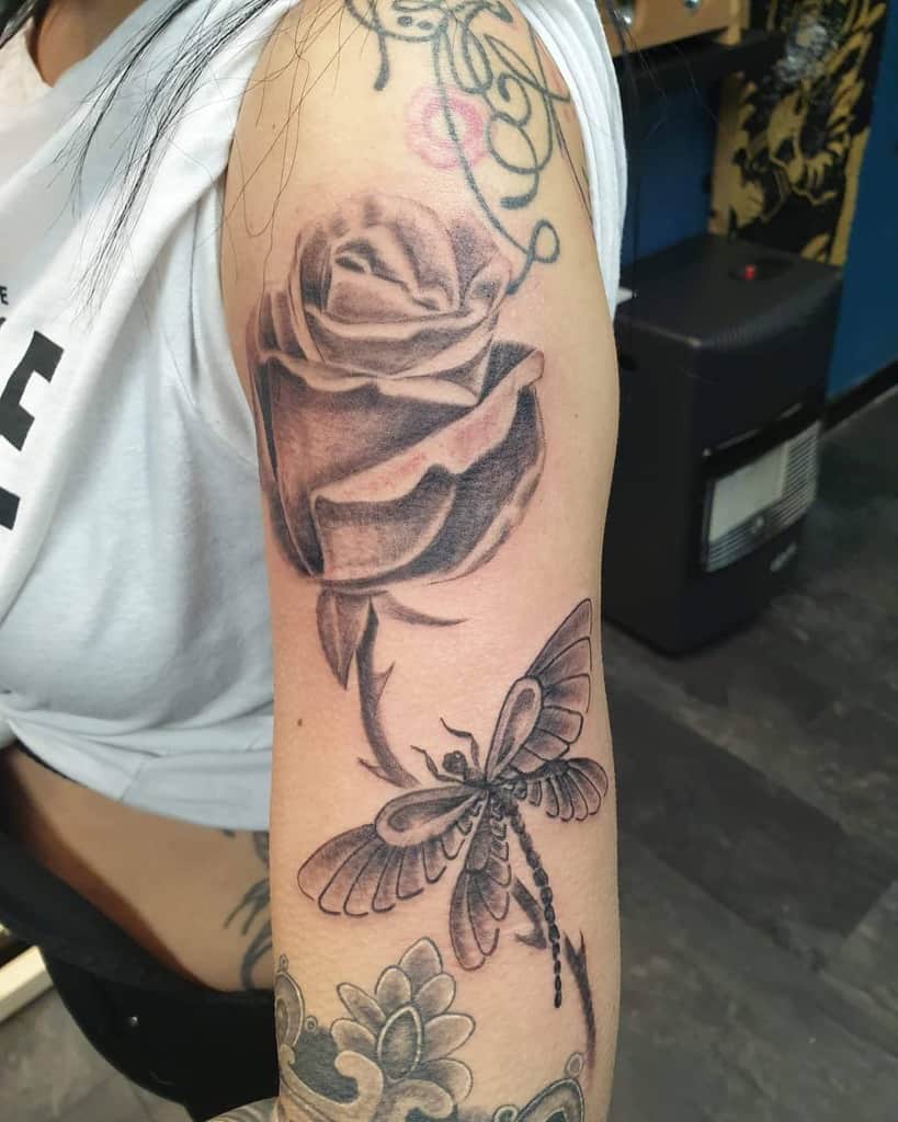 Black and grey tattoo featuring a detailed rose and a dragonfly on the upper arm, blending realism with soft shading.