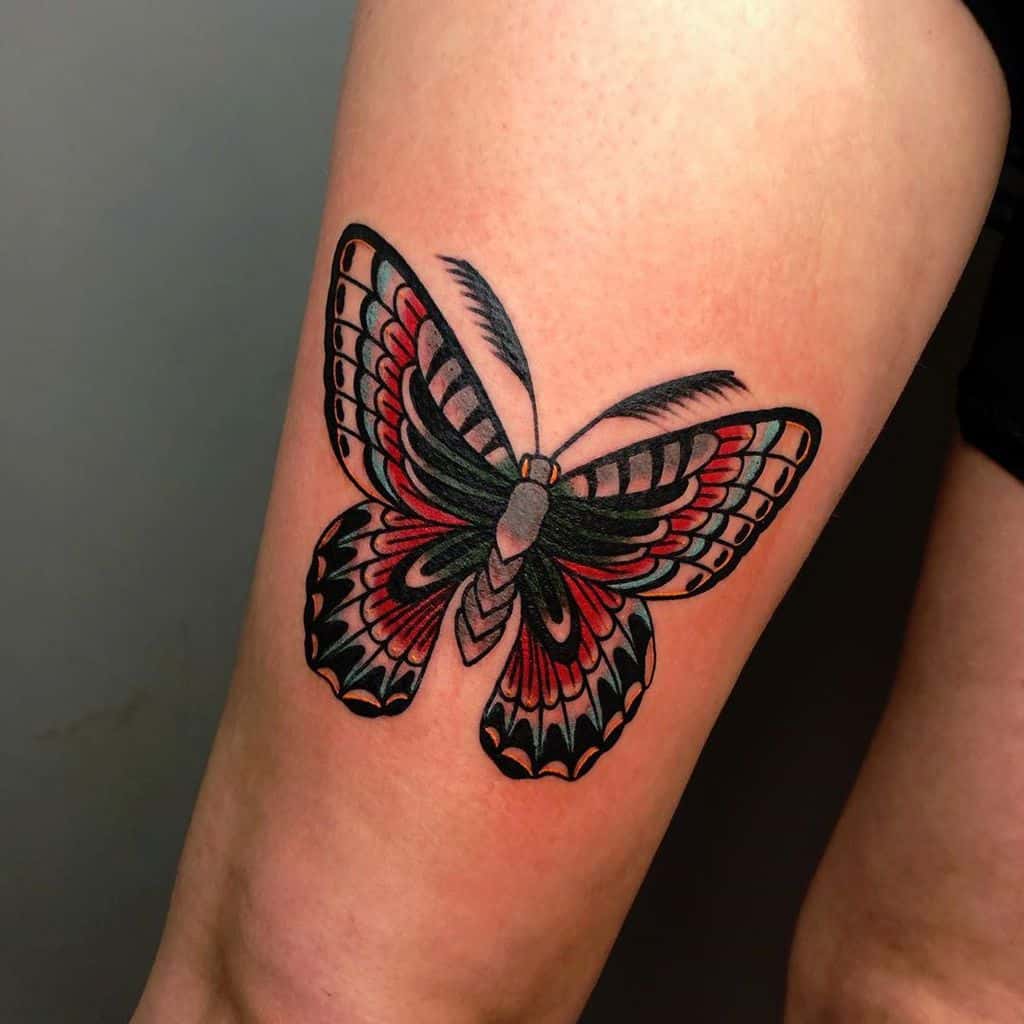 Traditional-style butterfly tattoo on the thigh, featuring bold black, red, and white shading with intricate wing details for a striking design.