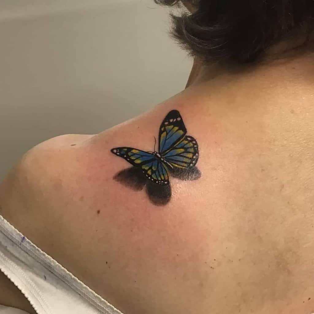 Realistic blue and yellow butterfly tattoo on the shoulder with a striking 3D shadow effect, creating a lifelike appearance.