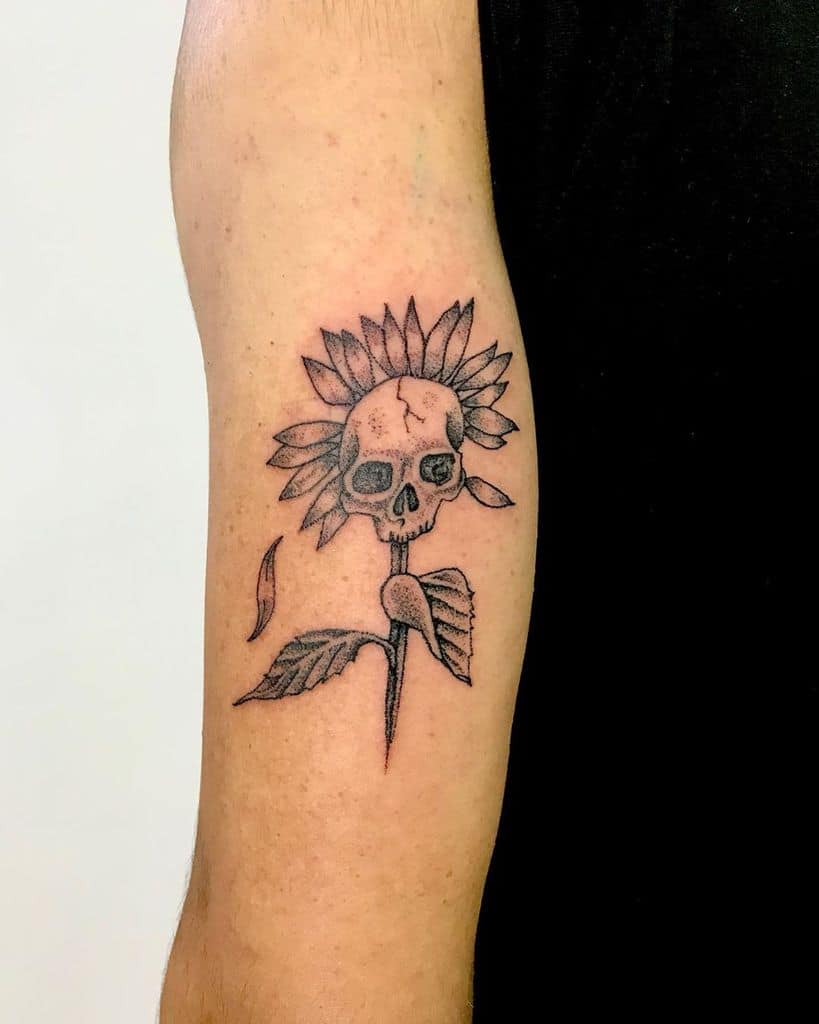 Black ink tattoo on upper arm featuring a sunflower with a skull at its center, fine line details, and subtle shading for a gothic floral design.