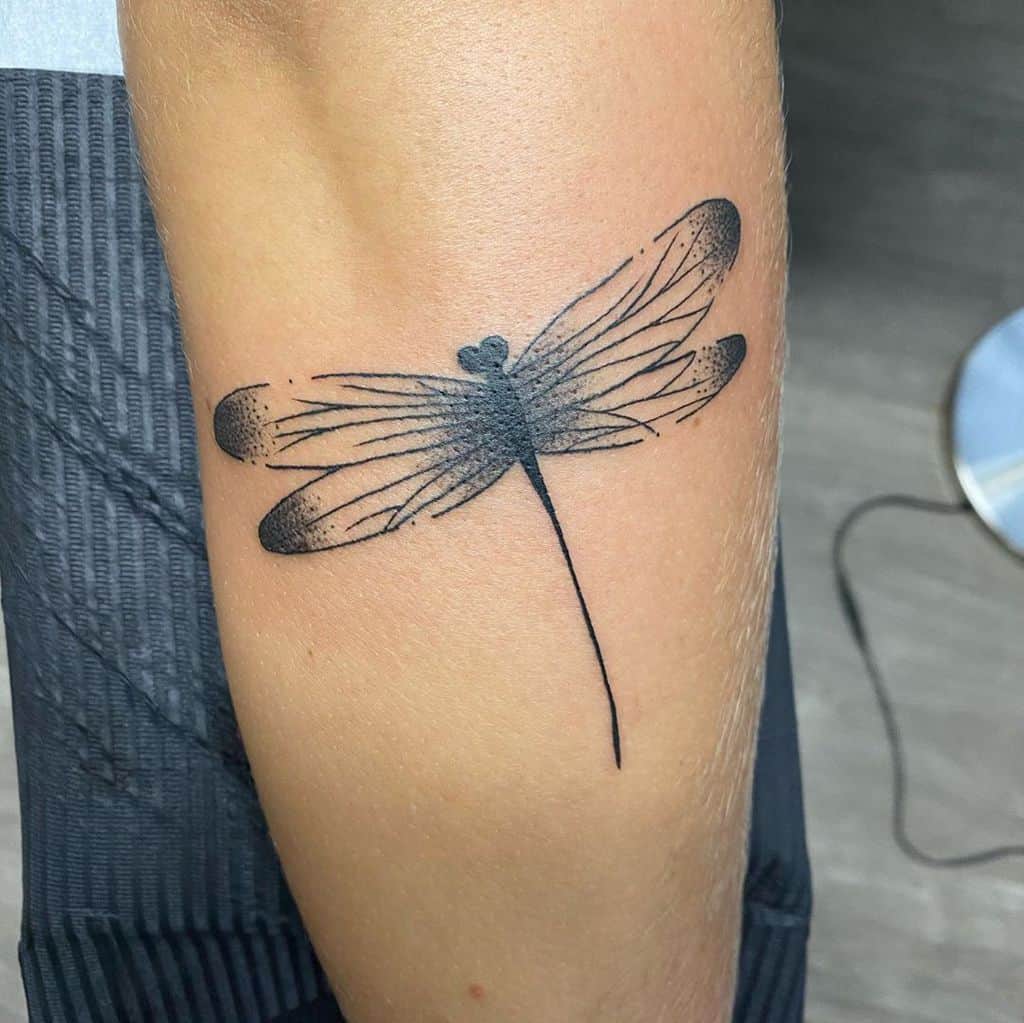 Minimalist black ink dragonfly tattoo on the arm, featuring fine lines and soft shading for an elegant, artistic effect.