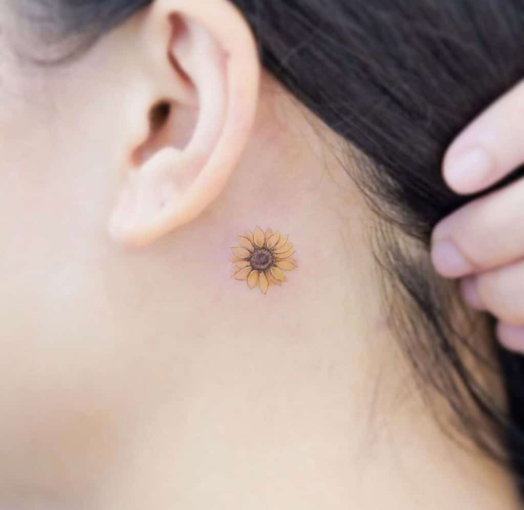 Small, delicate sunflower tattoo behind the ear with fine details, soft yellow petals, and a brown center for a subtle floral design.