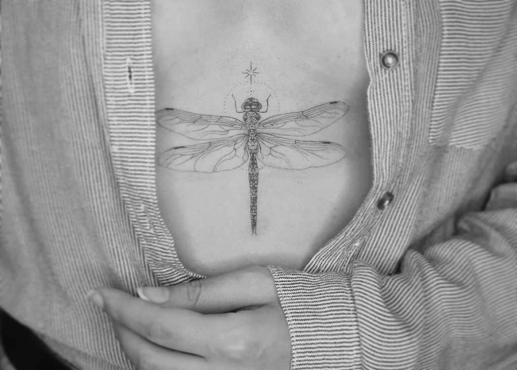 Elegant fine-line dragonfly tattoo on the sternum, featuring delicate details and a small celestial accent above.