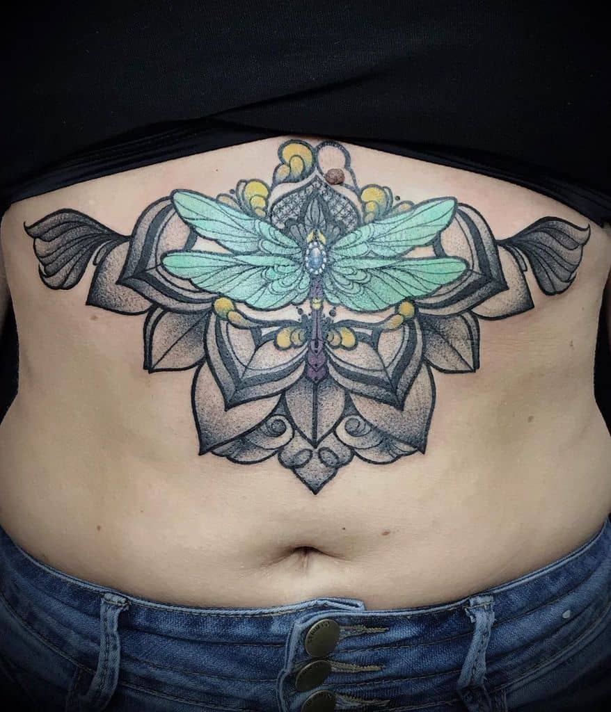 Intricate dragonfly tattoo on the lower abdomen, featuring mandala elements, bold black shading, and vibrant blue-green wings.