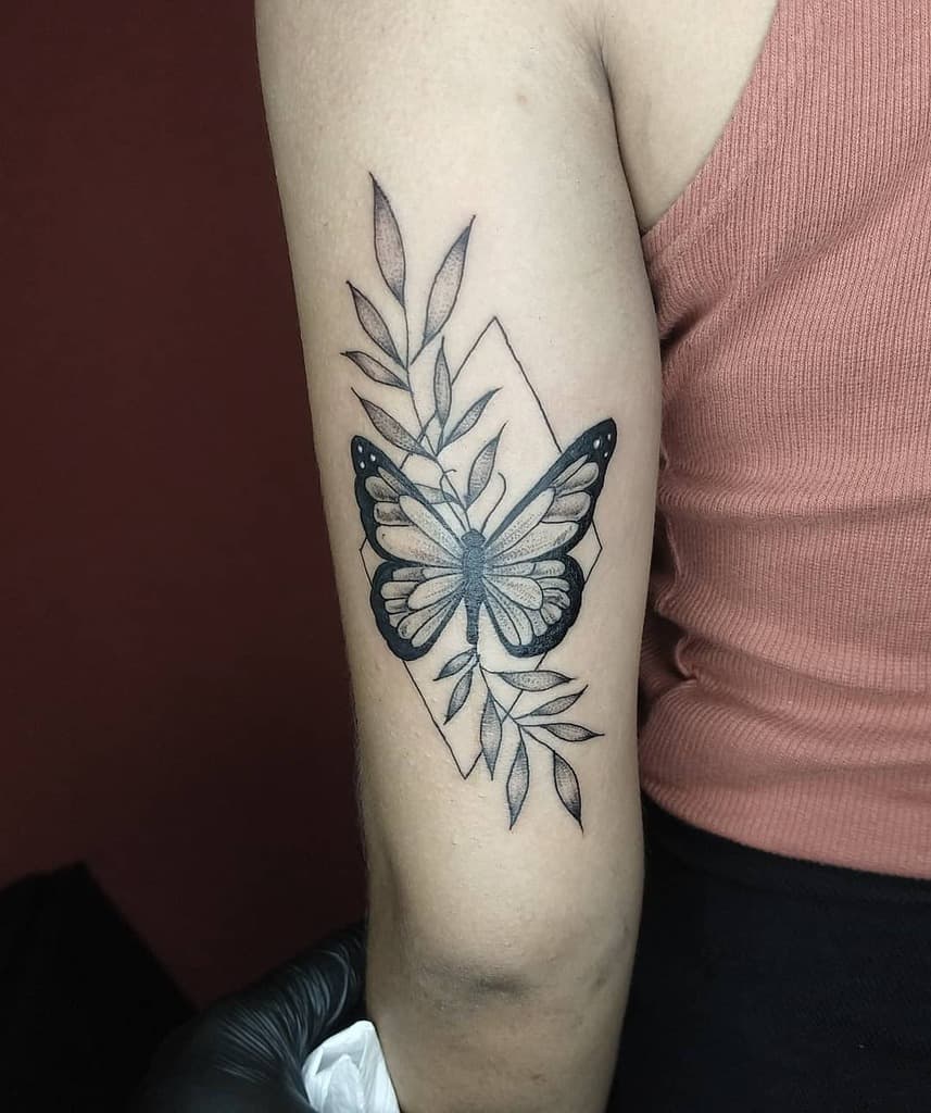 Elegant black and gray butterfly tattoo on the upper arm, featuring delicate leaves and a geometric outline for a modern, artistic look.